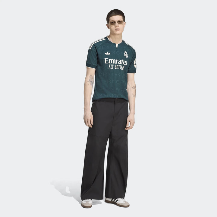 Male model wearing a dark green Real Madrid 26/27 Away Jersey, black wide-leg pants, and white sneakers, standing on a white