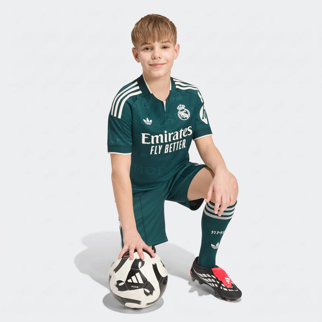 Young boy wearing a dark green Real Madrid 26/27 Away Kids Jersey kit, kneeling with a soccer ball.