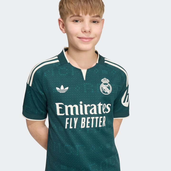 Smiling boy modeling a green Real Madrid 26/27 away jersey for kids, featuring Adidas stripes and sponsor logos.