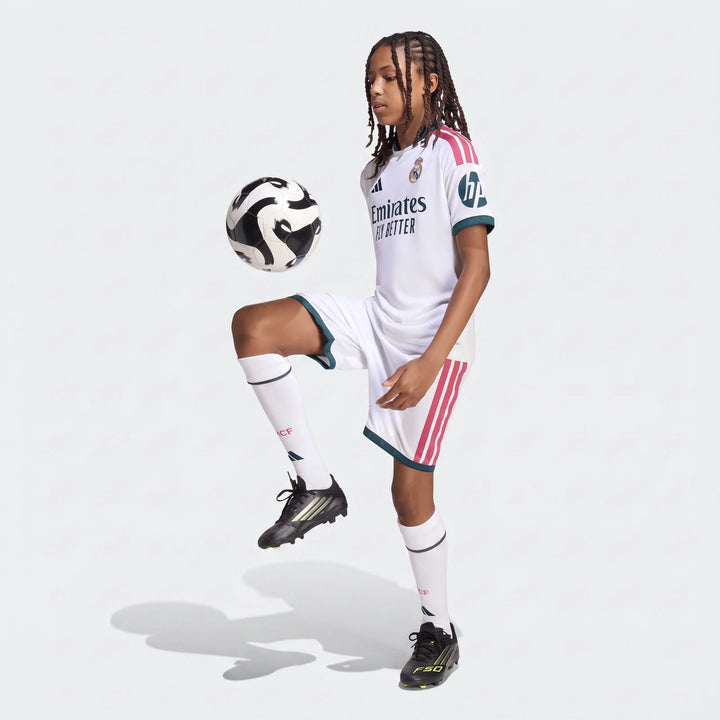 Young male model in a white Real Madrid 26/27 Home Jersey and shorts, juggling a soccer ball with black and yellow cleats.