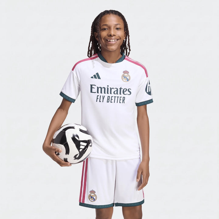 Young person wearing a white Real Madrid 26/27 Home Jersey and matching shorts, holding a soccer ball, smiling.