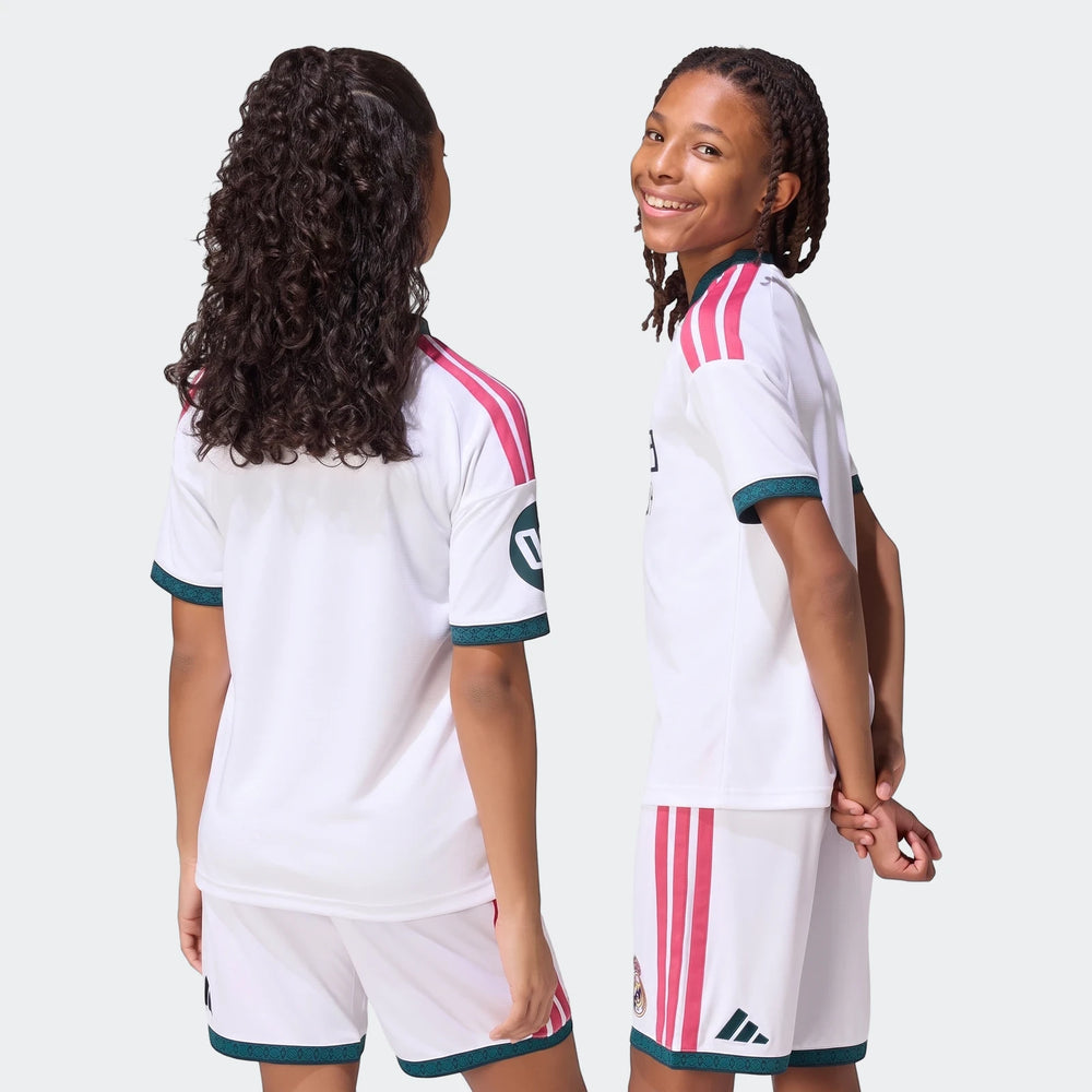 Two kids wearing white Real Madrid 26/27 Home Jerseys and matching shorts with pink stripes and green trim, one smiling.