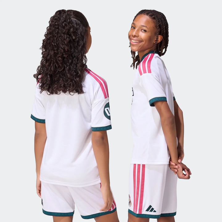 Two kids wearing white Real Madrid 26/27 Home Jerseys and matching shorts with pink stripes and green trim, one smiling.
