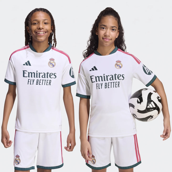 Two kids wearing white Real Madrid 26/27 home soccer jerseys and shorts, one holding a black and white soccer ball.