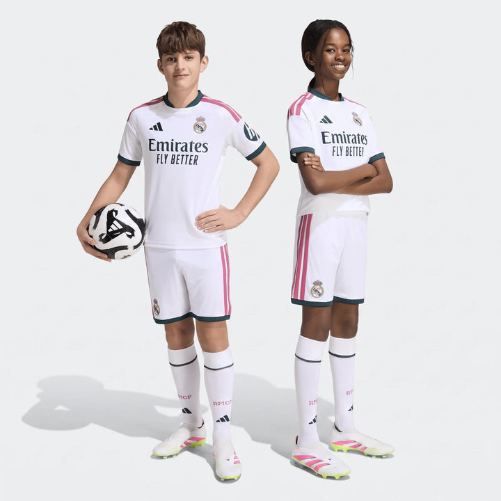 Two children wearing white Real Madrid 26/27 Home Shorts Kids and matching jerseys, one holding a football, posing in a