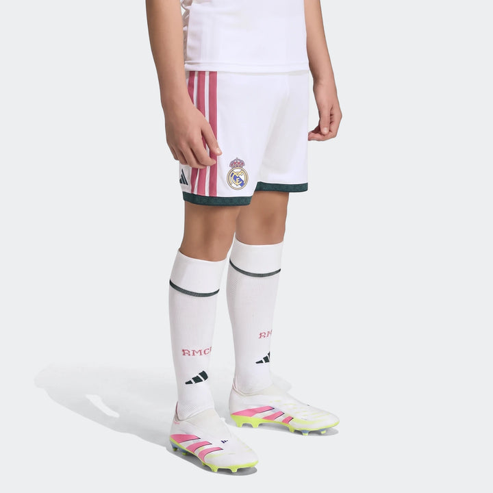 A child wears white Real Madrid 26/27 Home Shorts with maroon stripes and green trim, white socks, and white soccer cleats.