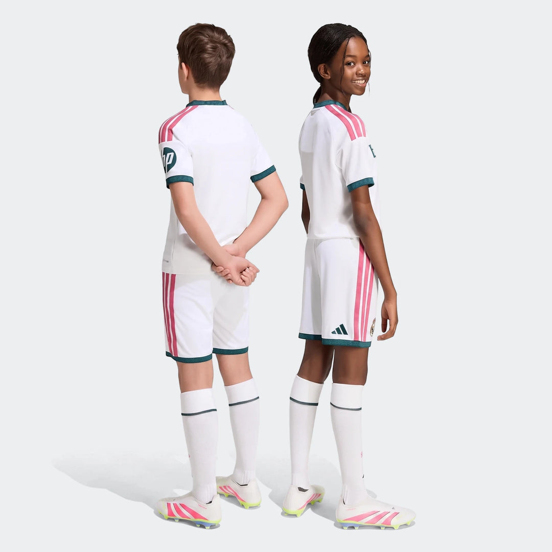 Two kids, a boy and a girl, modeling white Real Madrid 26/27 Home Shorts and matching jerseys with pink stripes and green