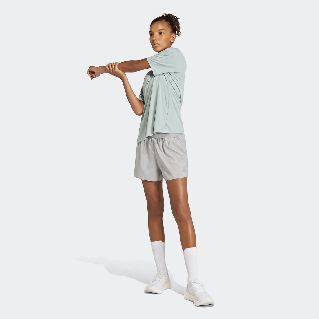 Young woman in mint green Run Essentials T-Shirt and grey shorts stretching, full body view.