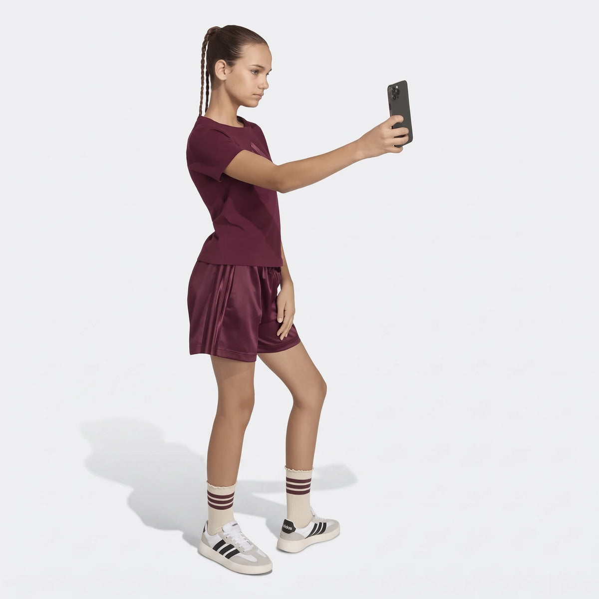 Young girl in a burgundy satin t-shirt and matching shorts, holding a black smartphone for a selfie.