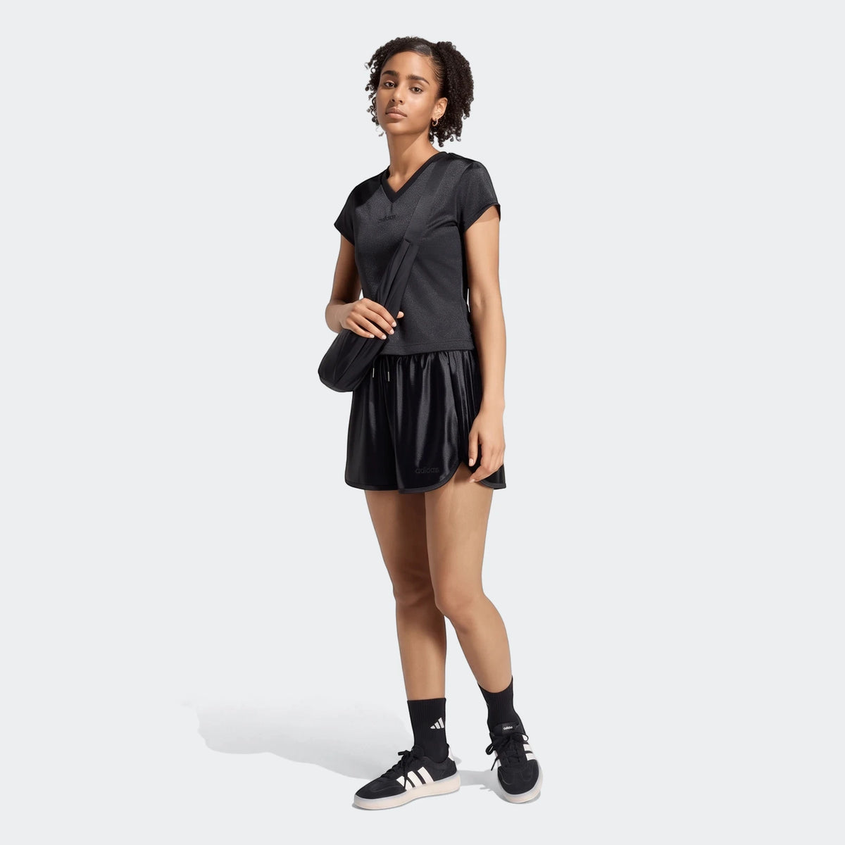 Full body shot of a female model wearing black Seasonal Essentials Satin Shorts, a black V-neck t-shirt, and black sneakers.