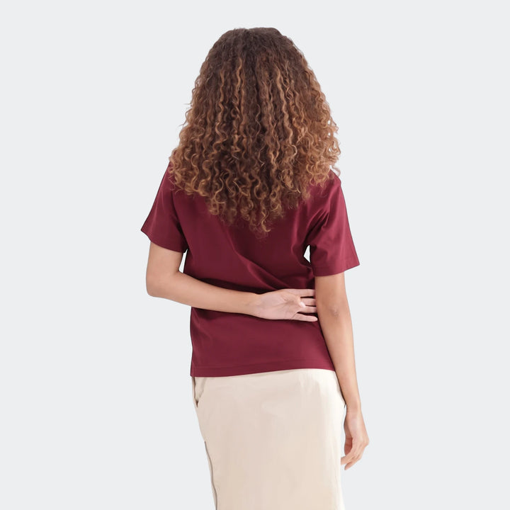Back view of a woman with curly hair wearing a maroon Slim Single Jersey T-Shirt and a light beige skirt, posed against a