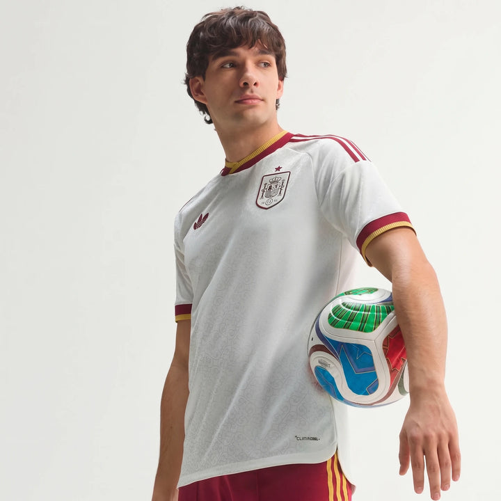 Spain 25/26 Authentic Away Jersey on a male model, white with red and gold trim, holding a multicolored soccer ball.