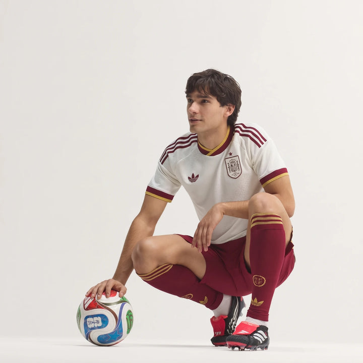 Male model wearing Spain 25/26 Authentic Away Jersey in white and maroon, crouching with a soccer ball.