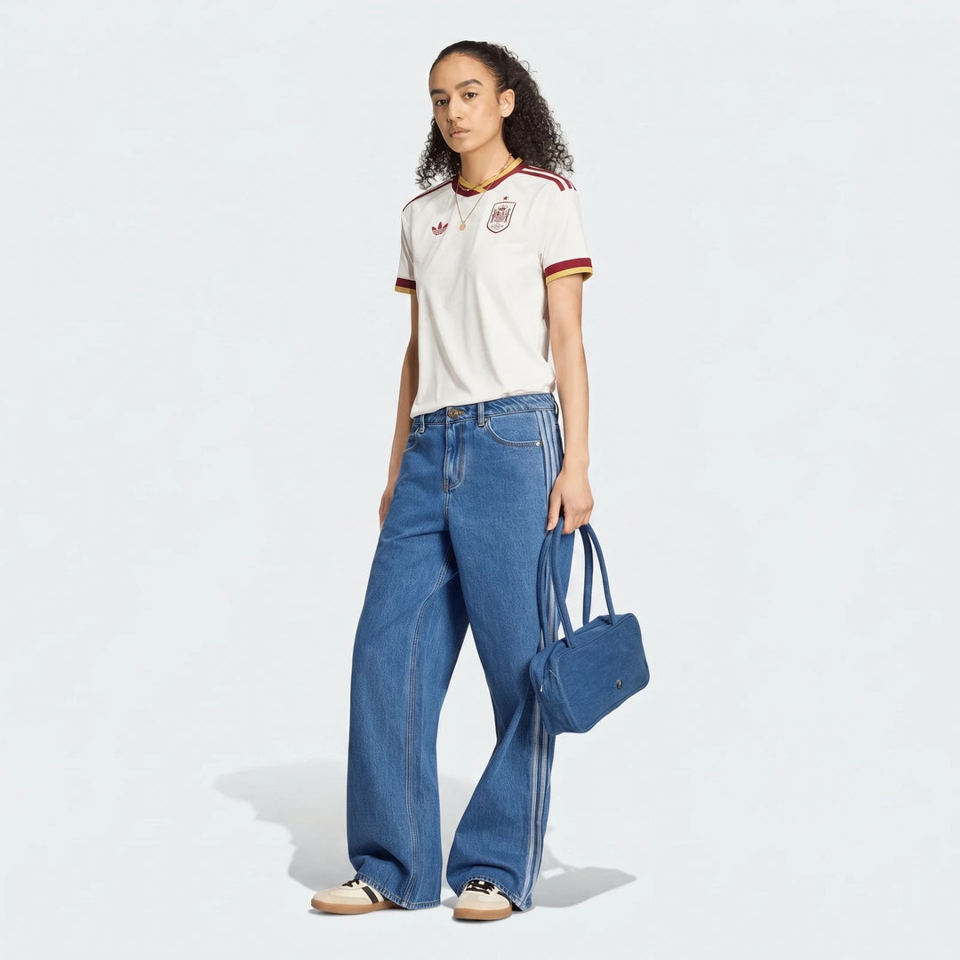 Women's Spain 25/26 Away Jersey in white with red and yellow details, worn with wide-leg denim jeans and holding a matching