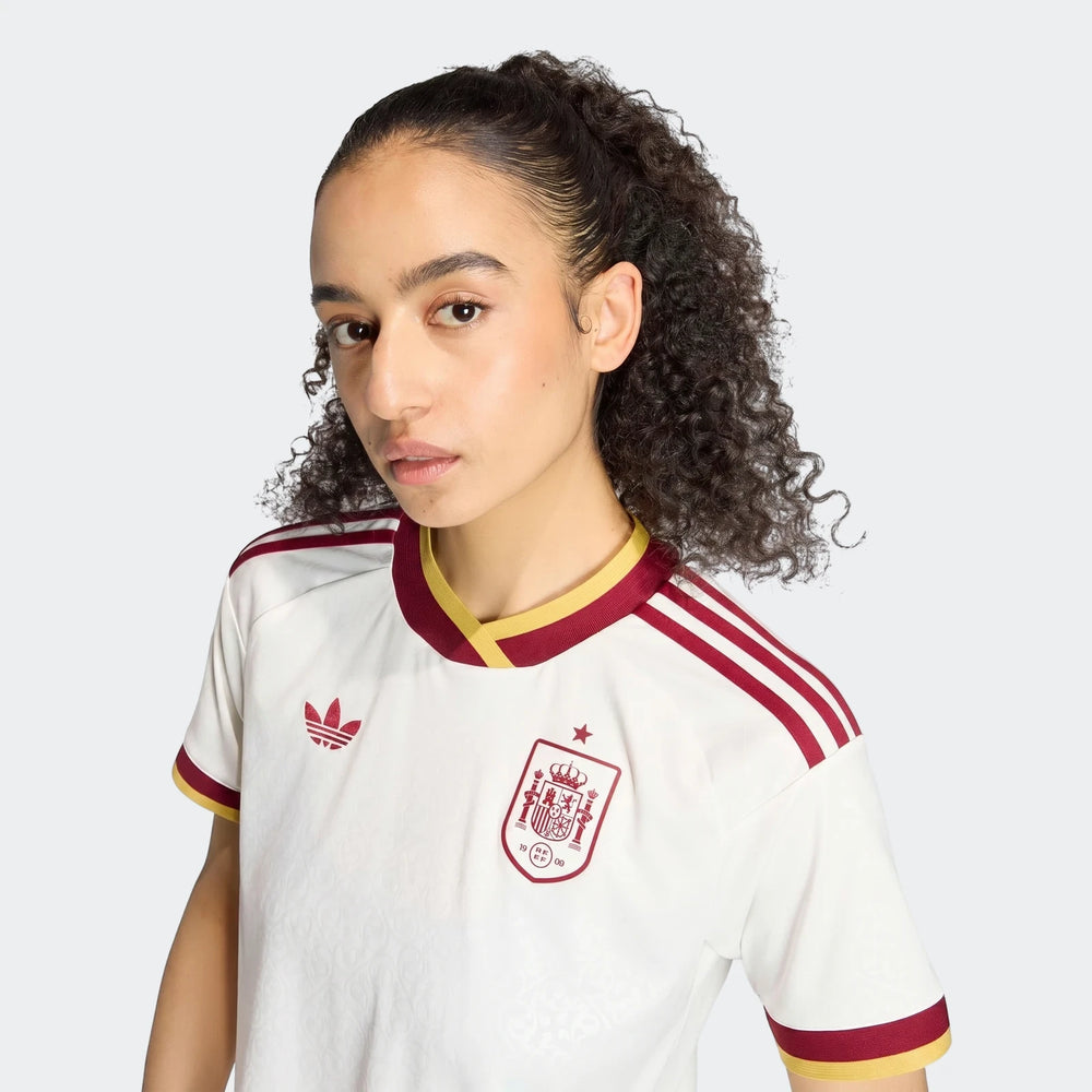 Front view of a woman wearing the white, red, and gold Spain 25/26 Away Jersey, featuring the team crest and Adidas logo.