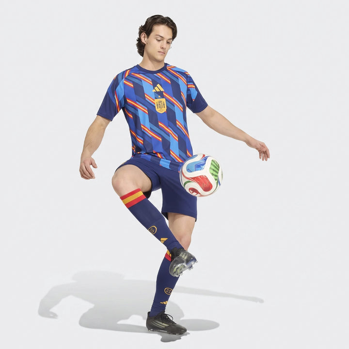 Male model in a blue patterned Spain 26 Home Pre Match Jersey and shorts, juggling a soccer ball, captured full length.