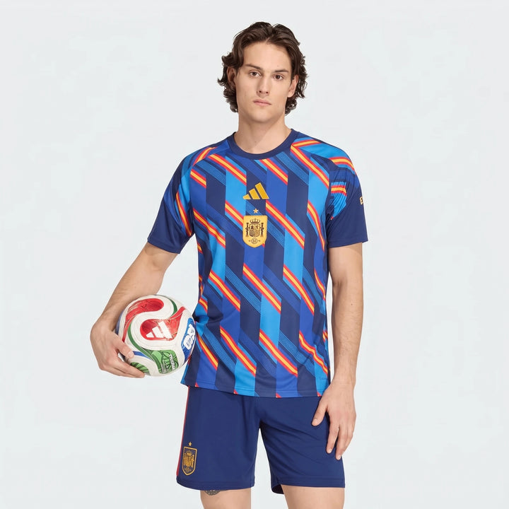 Male model wears the Spain 26 Home Pre Match Jersey and matching shorts, holding a soccer ball, in a studio setting.