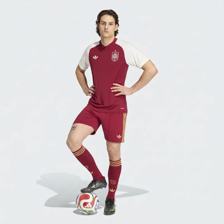 Spain Away Pre-Match Shirt worn by a male model, maroon with white sleeves and gold accents, standing with a soccer ball.