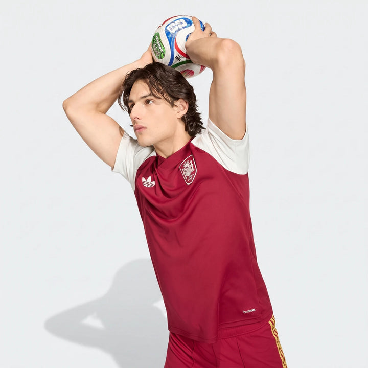 Male model wearing a red and white Adidas Spain Away Pre-Match Shirt, holding a multi-colored soccer ball above his head.