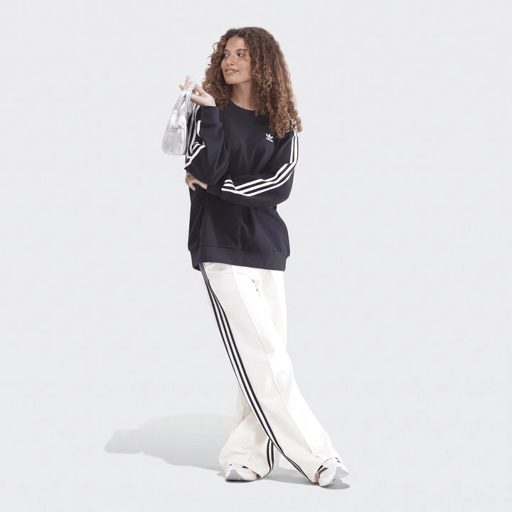 Woman wearing a black Adidas 3-Stripes oversized crew sweatshirt, white track pants, and holding a small silver handbag.