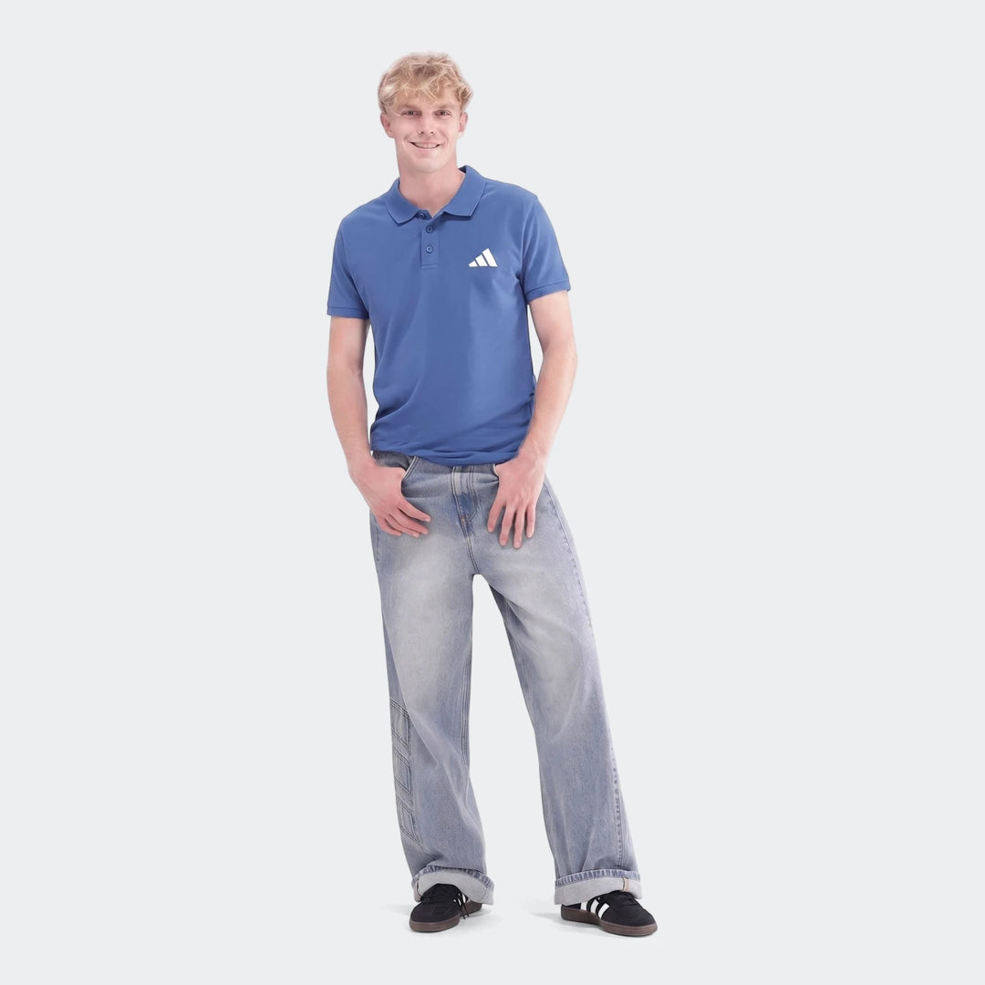 A man stands full body wearing a blue Sportswear Slim Pique Polo Shirt, light wash jeans, and black sneakers. The polo shirt