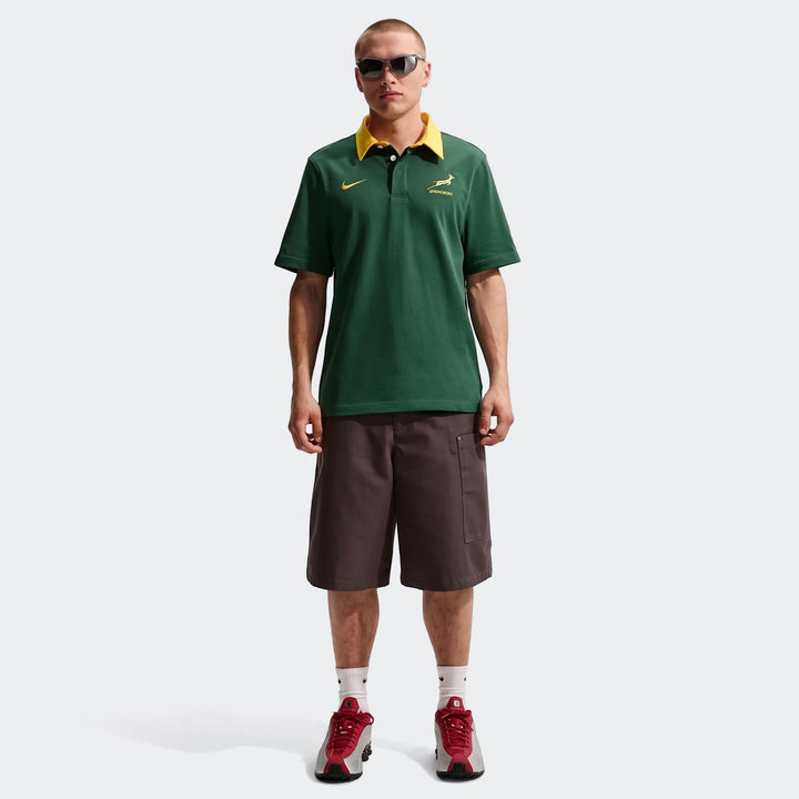 Man wearing a green Springboks Men'S Rugby Home Short-Sleeve Jersey with dark brown shorts and red sneakers.