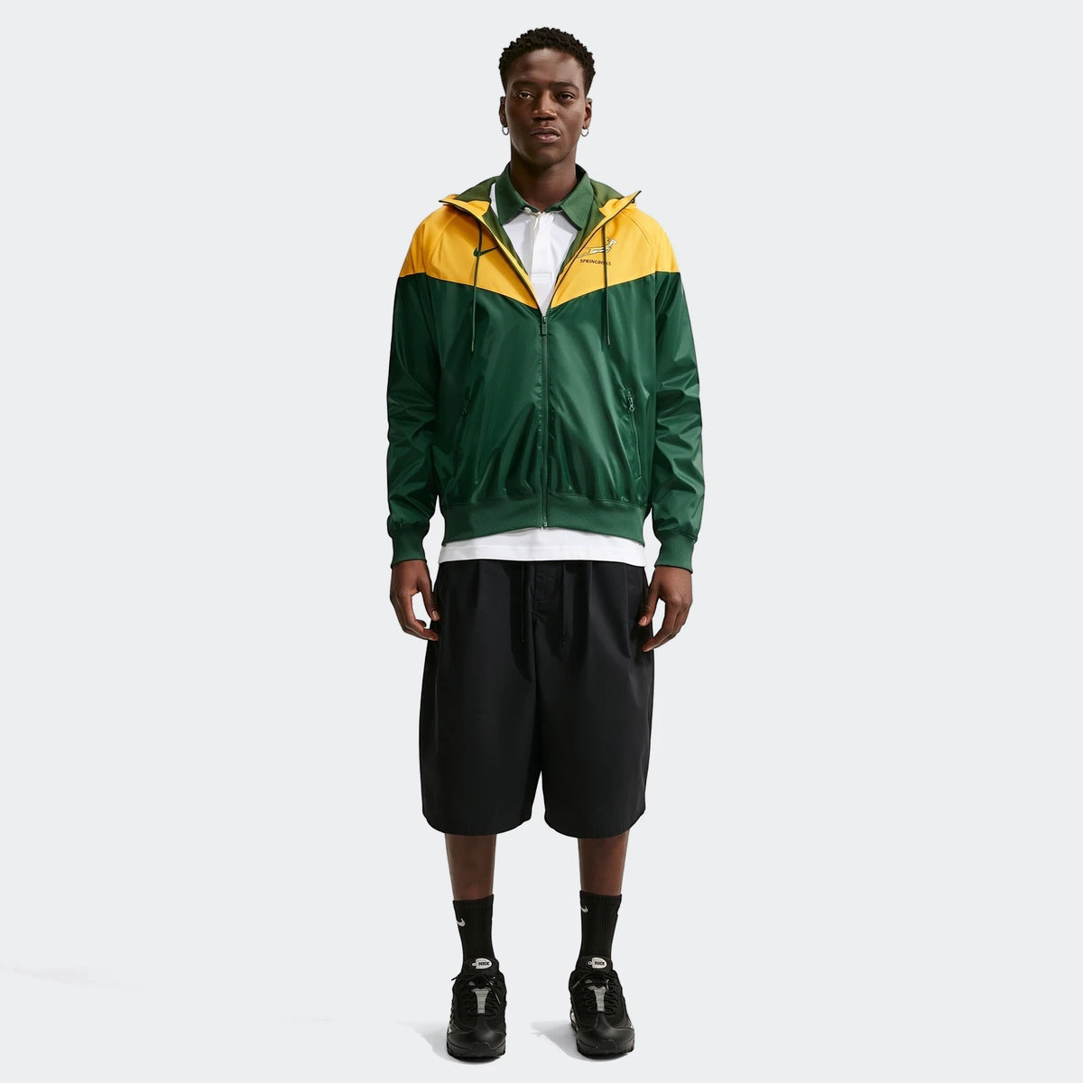 Full body shot of a male model wearing a green and yellow Springboks Nike Windrunner jacket with black shorts and sneakers.