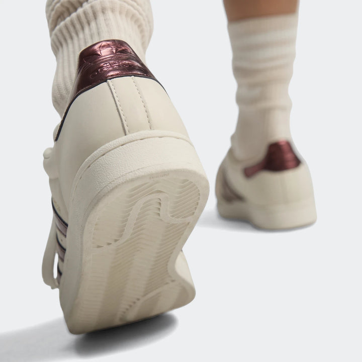 Cream Adidas Superstar II shoes with metallic reddish-brown heel tab and visible sole, worn with ribbed socks, shown from a