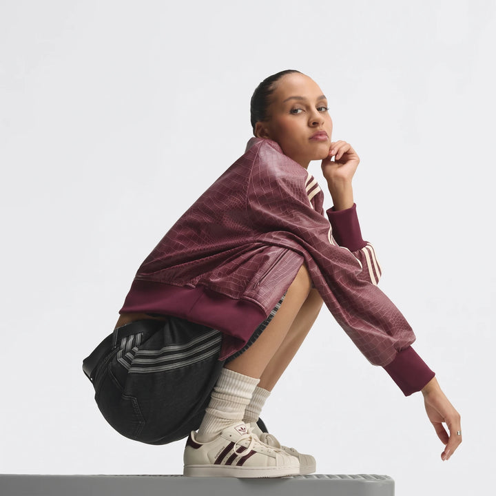 Woman wearing maroon crocodile-textured bomber jacket, dark denim shorts, and cream Adidas Superstar II shoes, squatting on
