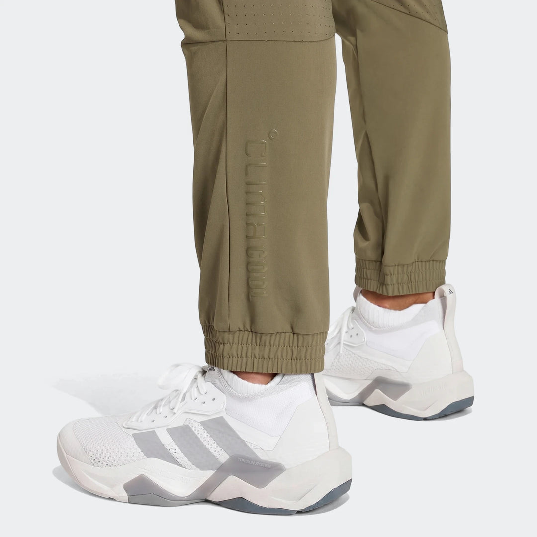 Close-up of olive green Technical Apparel Slim Pants with embossed logo and ribbed cuffs, worn with white sneakers.