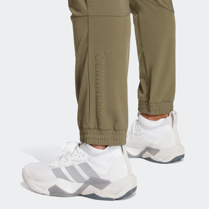 Close-up of olive green Technical Apparel Slim Pants with embossed logo and ribbed cuffs, worn with white sneakers.
