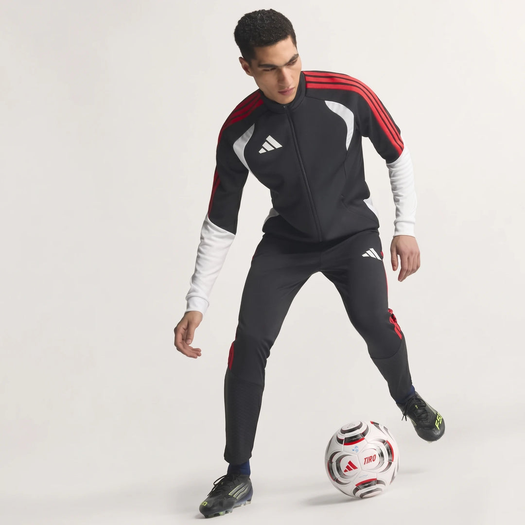Male model in black, red, and white Adidas Tiro 26 Competition Training Jacket and matching pants, dribbling a soccer ball.