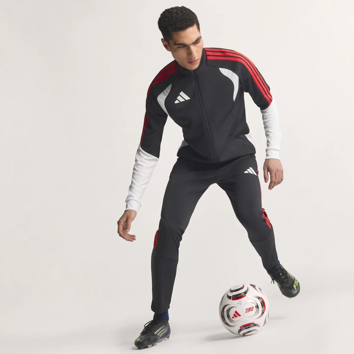 Male model in black, red, and white Adidas Tiro 26 Competition Training Jacket and matching pants, dribbling a soccer ball.