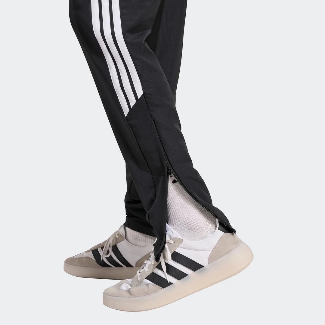 Black Adidas Tiro 26 League Presentation Pants with white stripes, worn by a person with sneakers, shown in a side view.