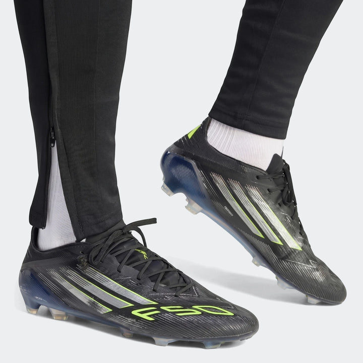 Close-up of a person wearing black Tiro 26 League Training Tracksuit Bottoms with zippers and black and neon green Adidas