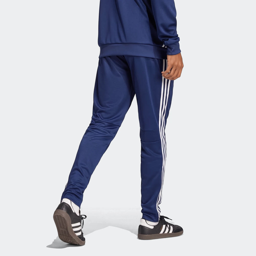 Male model wearing blue Adidas Tiro Essentials Pants with white stripes and black sneakers, viewed from behind.