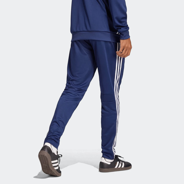 Male model wearing blue Adidas Tiro Essentials Pants with white stripes and black sneakers, viewed from behind.