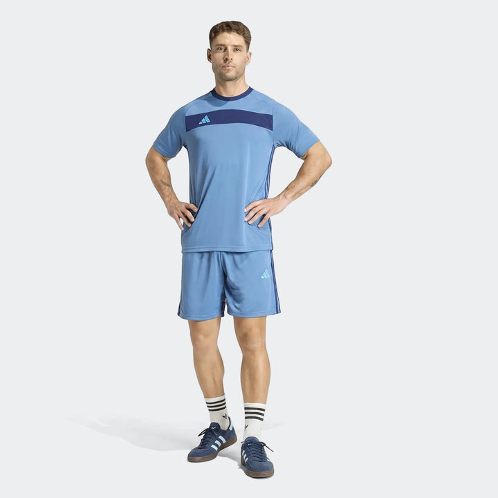 Male model wearing blue Adidas Tiro Essentials shorts and matching t-shirt, white striped socks, and navy sneakers, standing