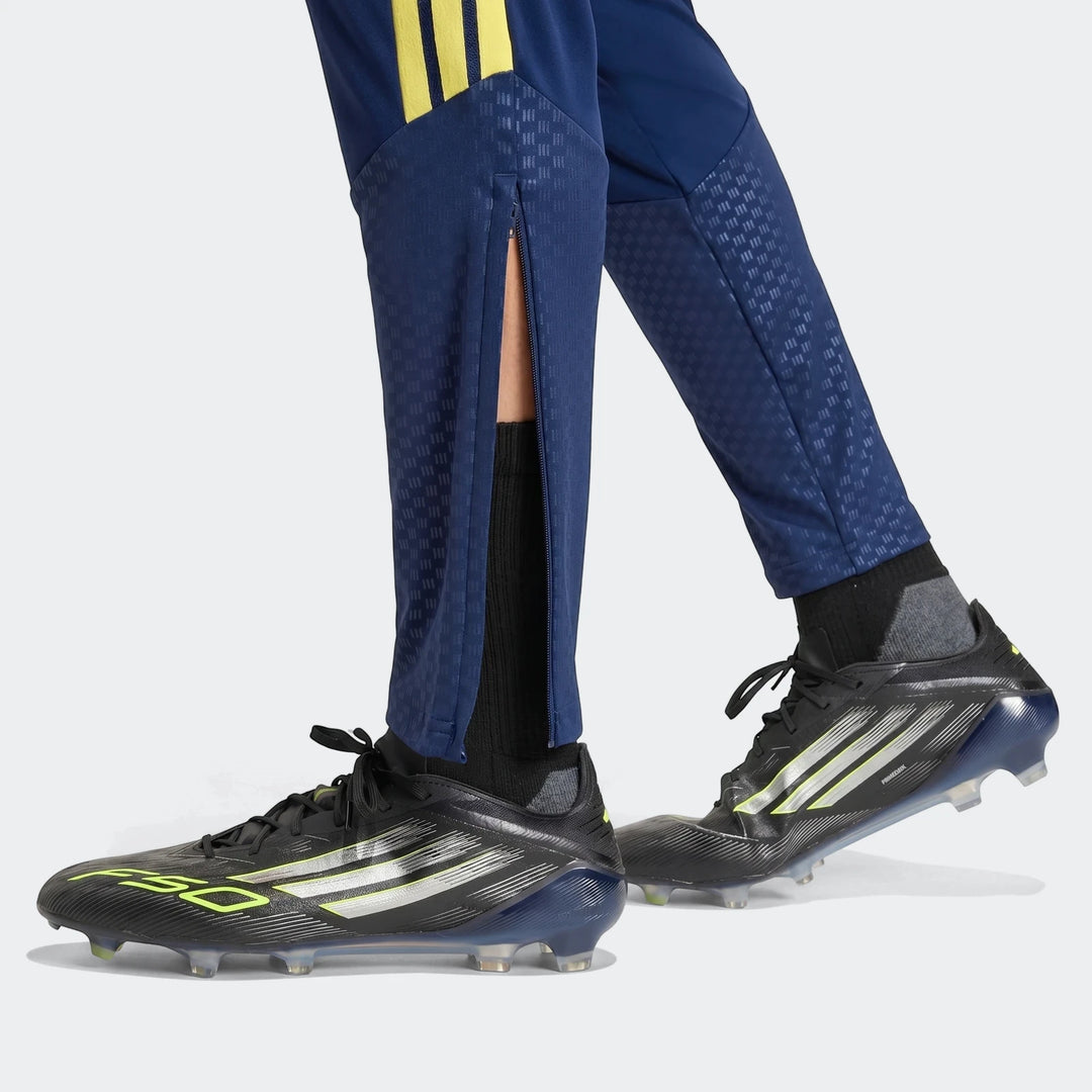 Tiro26 C Pant Blue Pants with yellow stripes and textured panels on a person's lower legs wearing black soccer cleats.