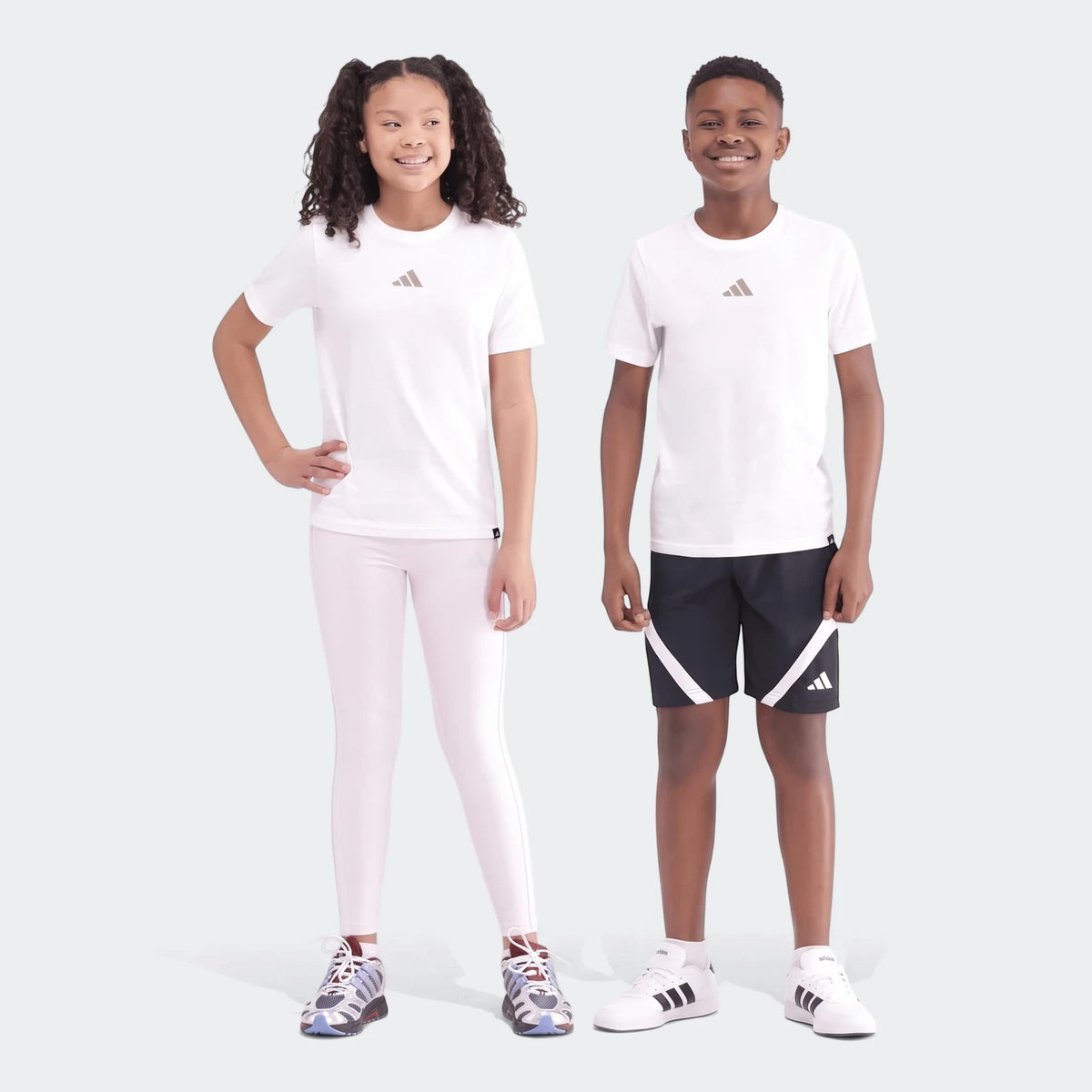 Two smiling children, a girl and a boy, wearing white Camo Graphic Tees with grey logos, shown full body.