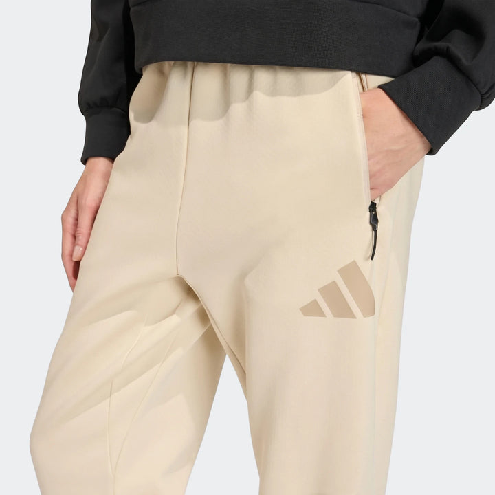 Close-up of a person wearing beige Adidas W Z.N.E. Pt trackpants with a black top, featuring a zippered pocket and Adidas