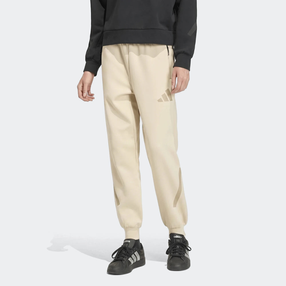 Beige Adidas Z.N.E. track pants with tonal logo, worn by a person with black Adidas sneakers. Studio shot.
