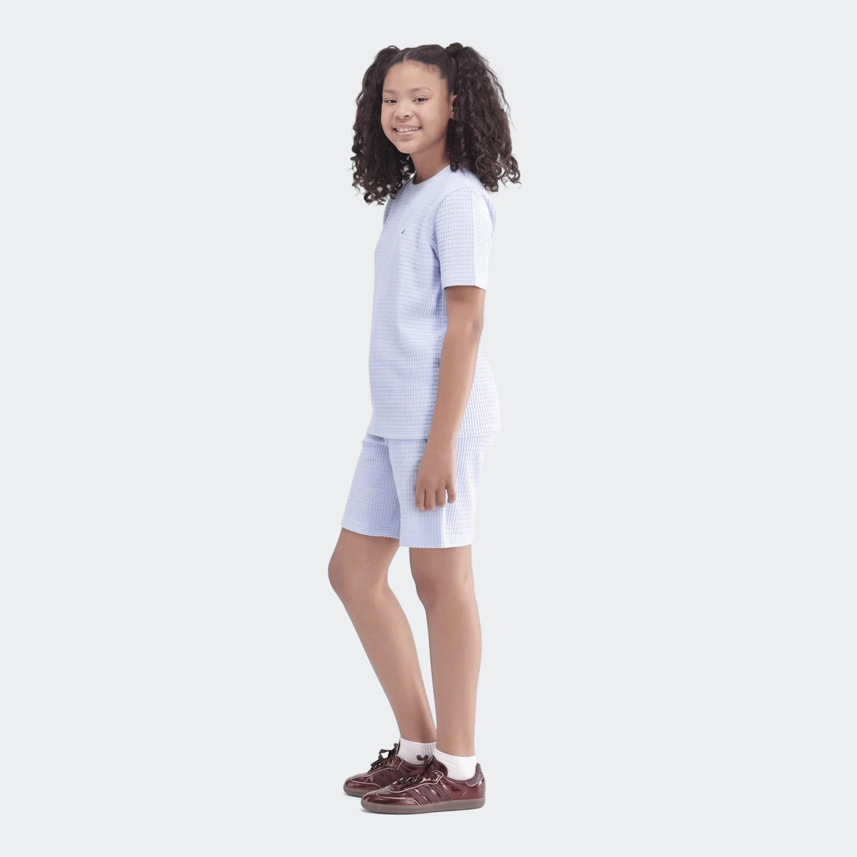 Young girl smiling in a light blue waffle knit short set, featuring a t-shirt and shorts, with shiny brown sneakers.