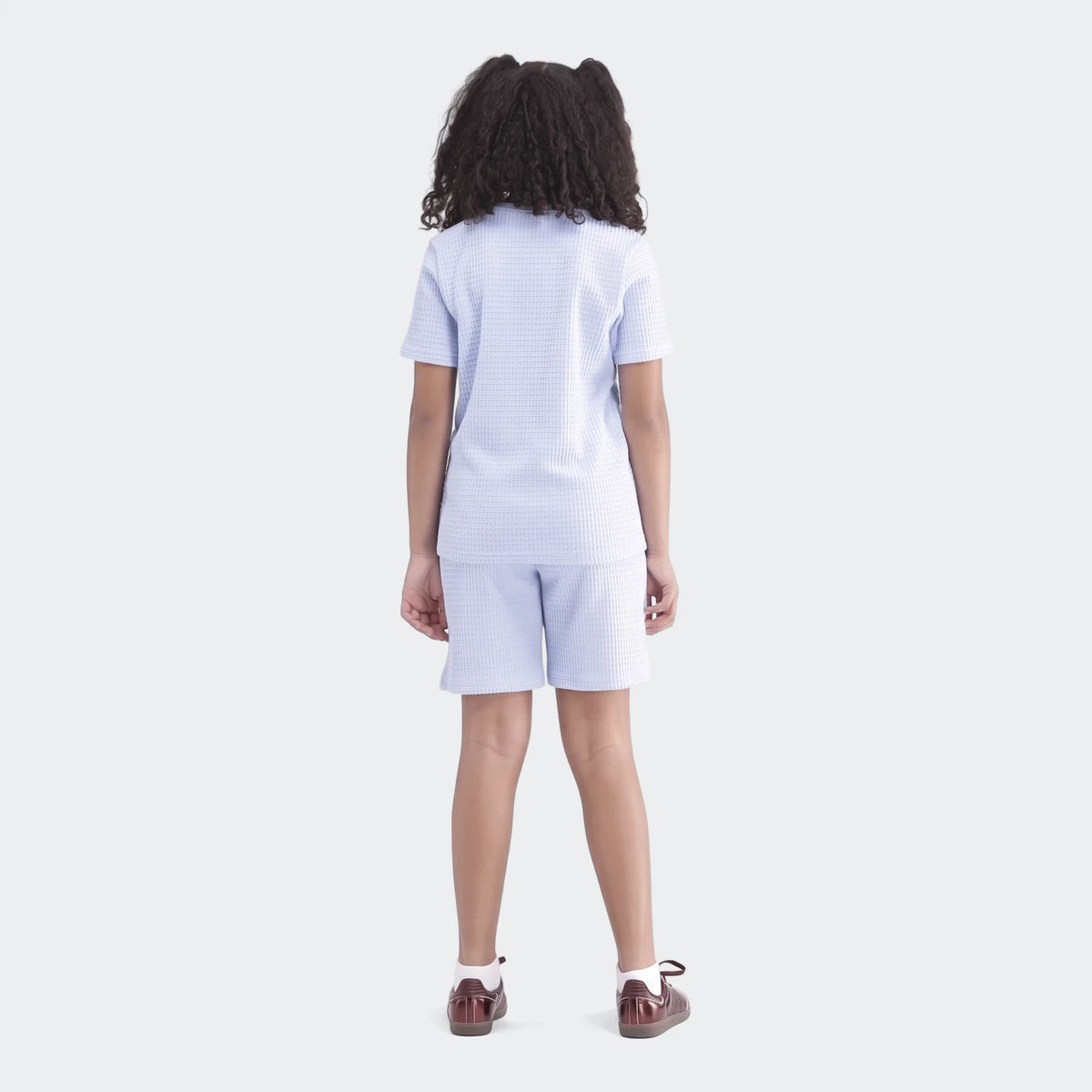 Young girl in a light blue waffle knit short-sleeve top and matching shorts, seen from the back, wearing brown shoes.