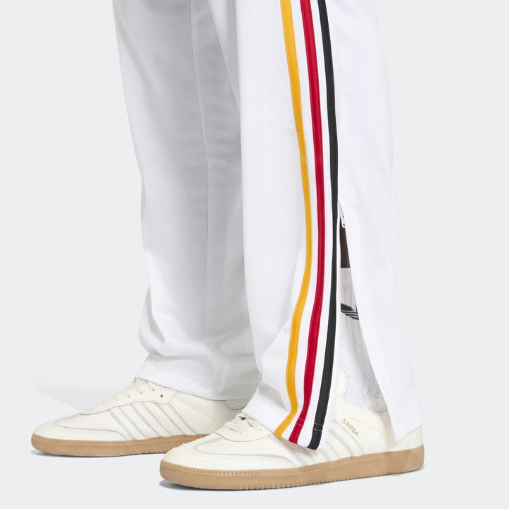Close-up of white Firebird Track Pants with red, yellow, and black stripes, worn with cream Adidas Samba sneakers.