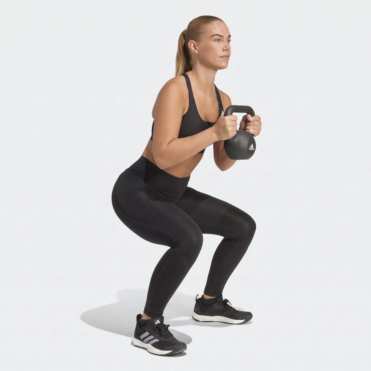 Woman in black Adidas sports bra and leggings performing a kettlebell squat. She holds a black kettlebell with both hands.