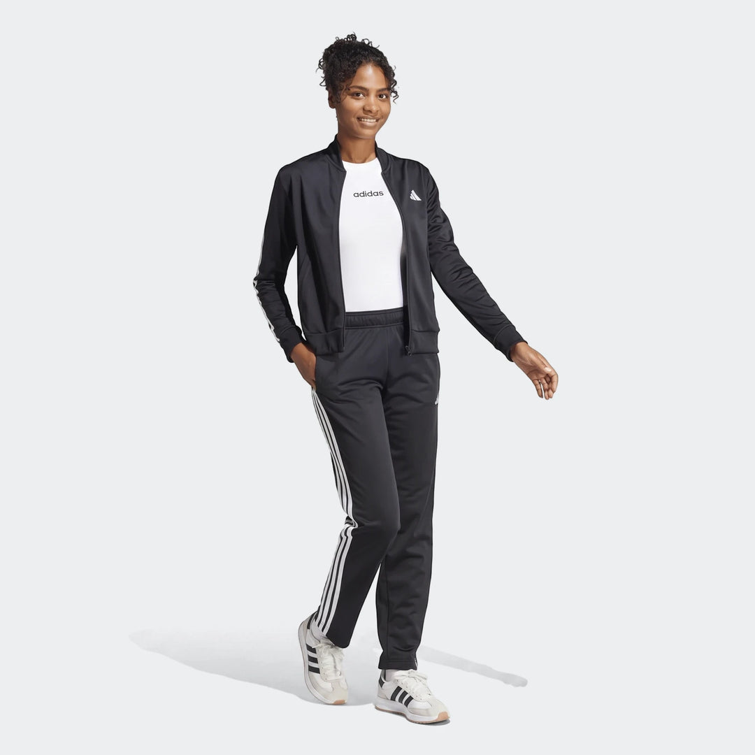Woman models black Adidas Dayready tracksuit with white stripes and white t-shirt, standing full-body in athletic sneakers.