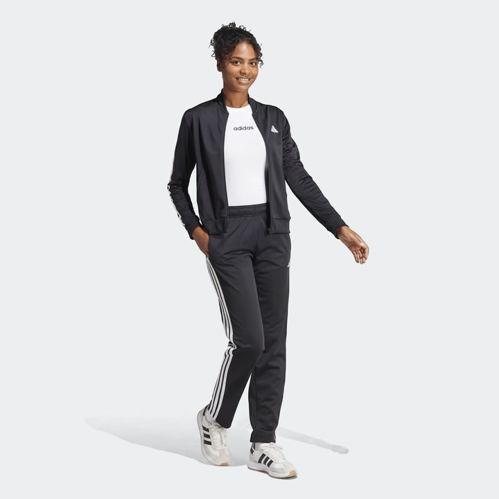 Woman models black Adidas Dayready tracksuit with white stripes and white t-shirt, standing full-body in athletic sneakers.