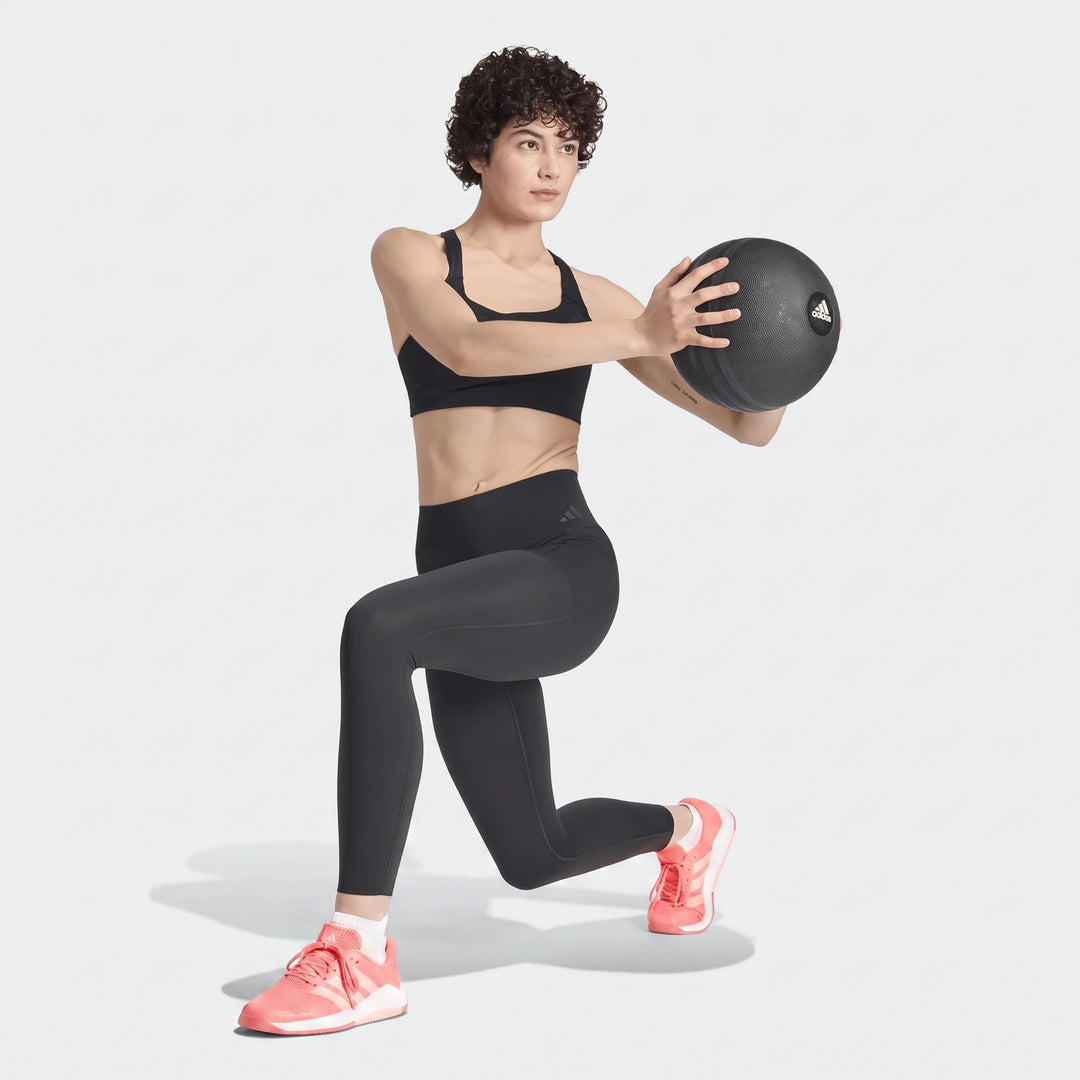 Woman in black Adidas Optime Workout Contour Full Length Leggings, black sports bra, and peach shoes in a lunge with a