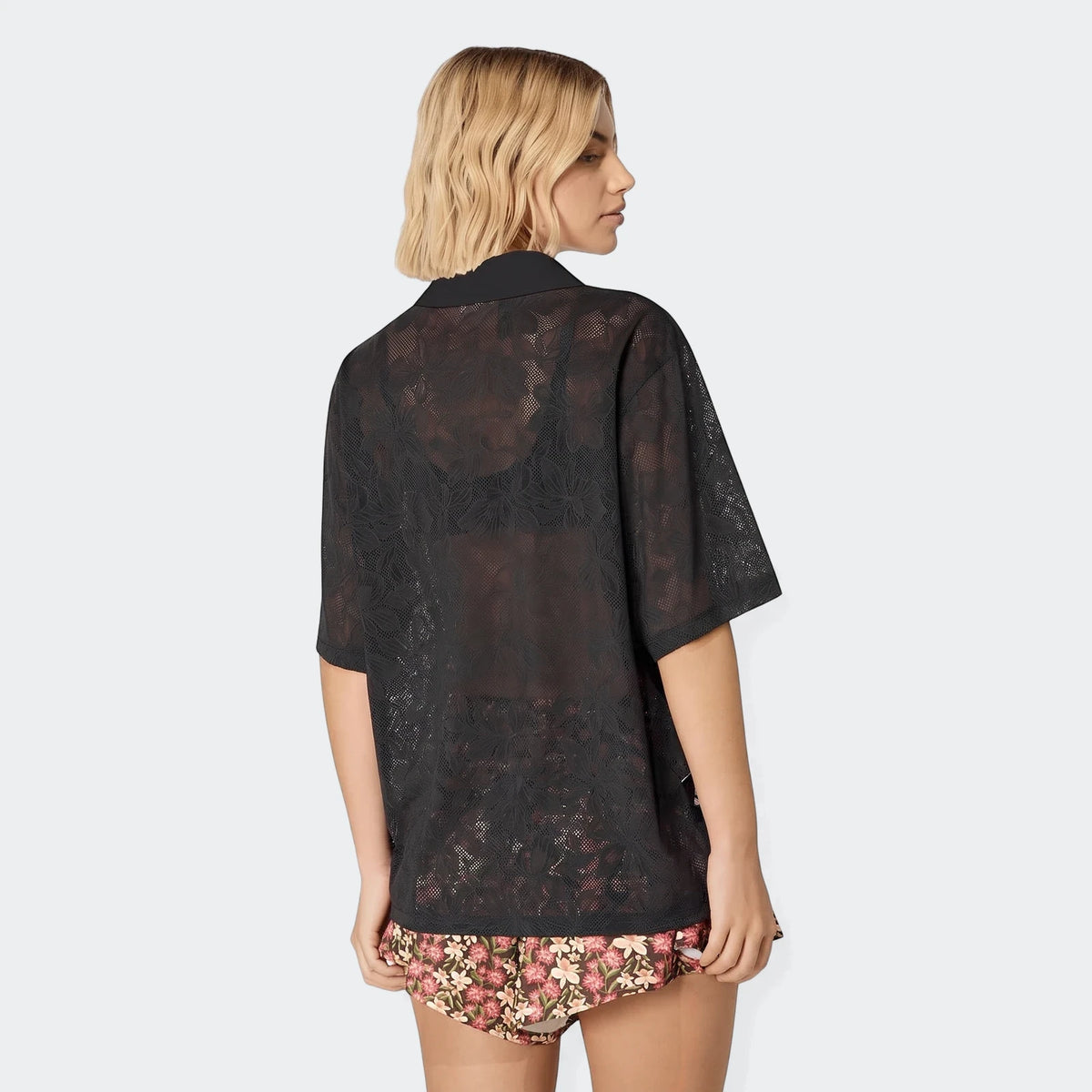 Woman wearing a black sheer lace collared shirt over a black bra and floral patterned shorts, profile view.